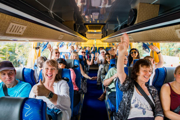 A front-view shot of a large multi-ethnic group of tourists celebrating on a coach bus, they are smiling and raising their arms with excitement, they are ready to begin their journey.